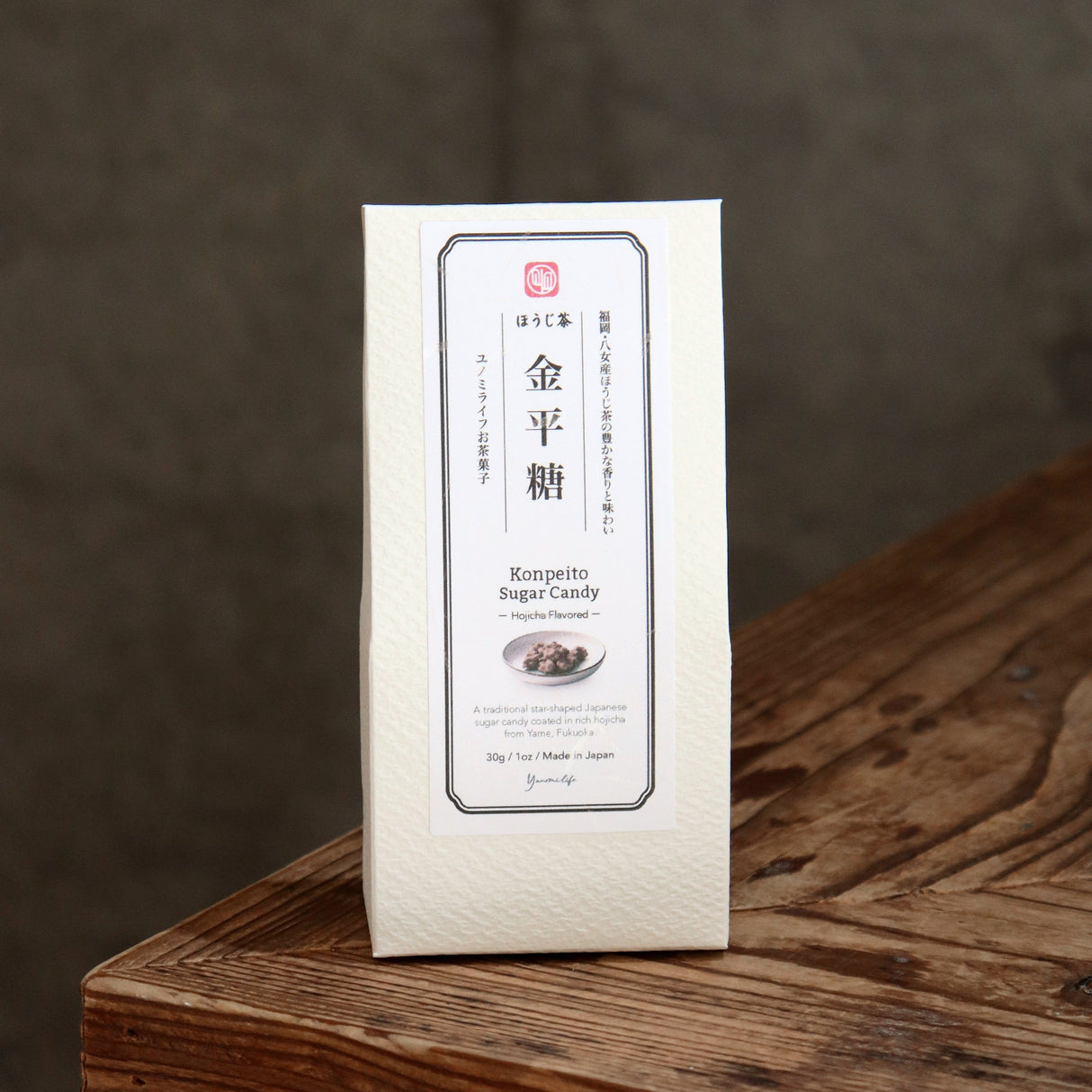 A white rectangular package of Japanese konpeito sugar candy stands upright on a rustic wooden surface against a dark gray background.