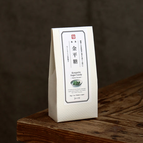 A white paper bag of Japanese Kompetto Sugar Candy standing on a rustic wooden surface against a dark brown background.